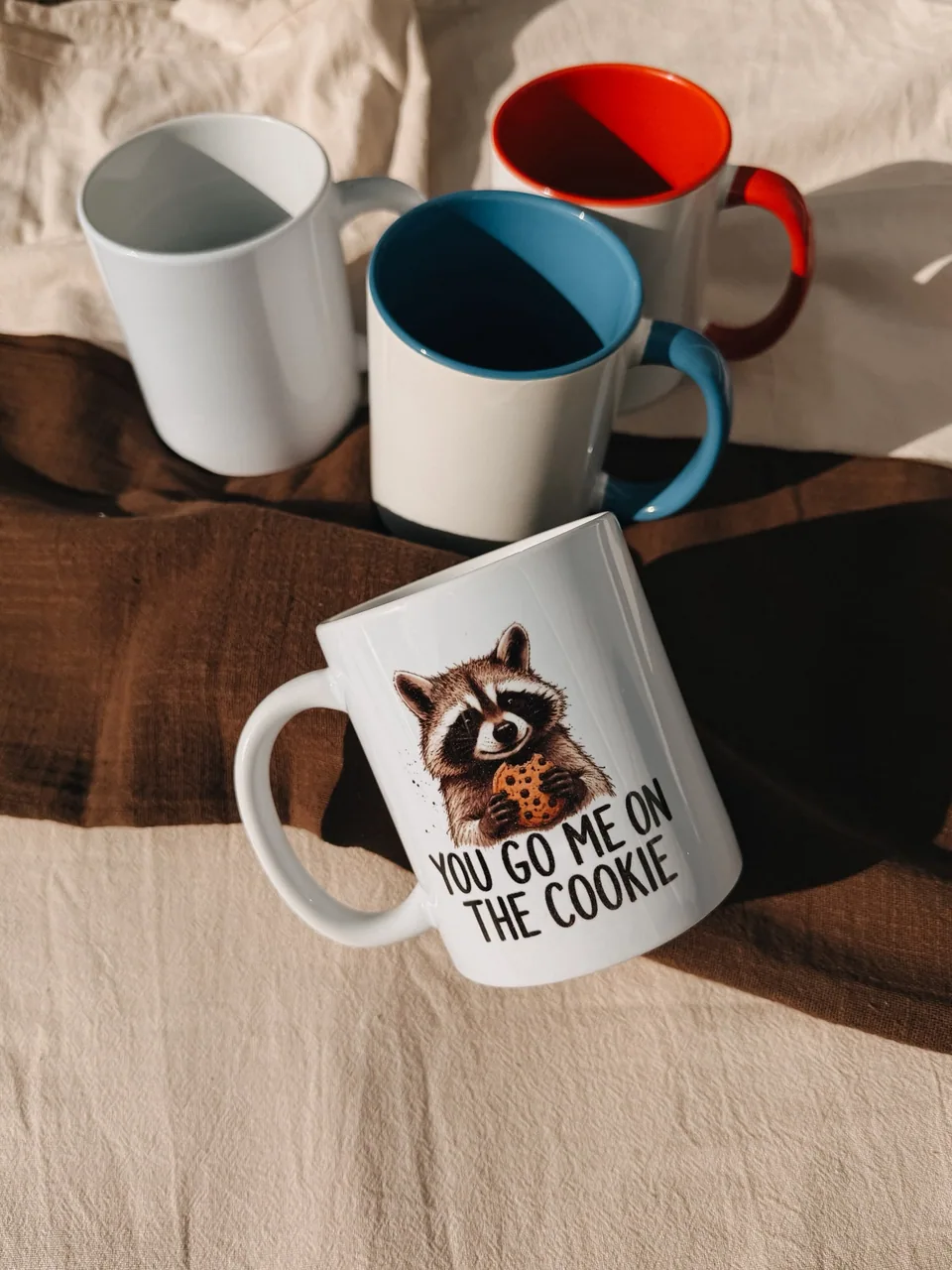 Drei weiße Tassen mit unterschiedlich gefärbten Henkeln hinter einer Tasse mit Waschbär-Aufdruck